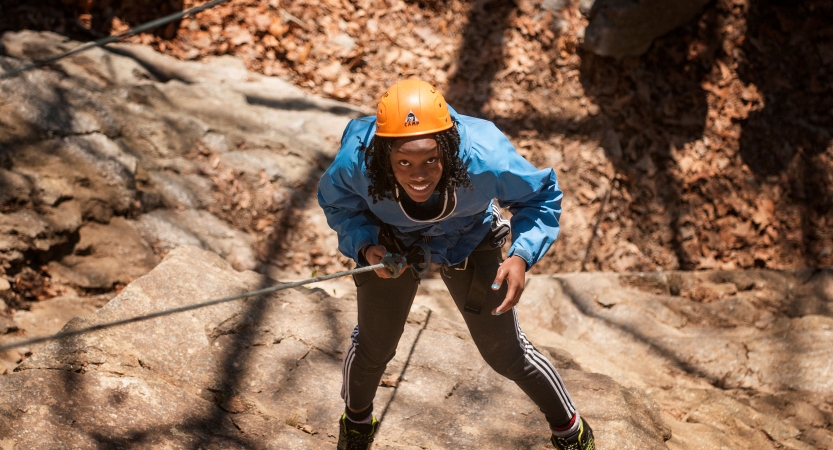 A young person wearing safety gear is secured by ropes as they look up toward the camera and smile while rock climbing. 